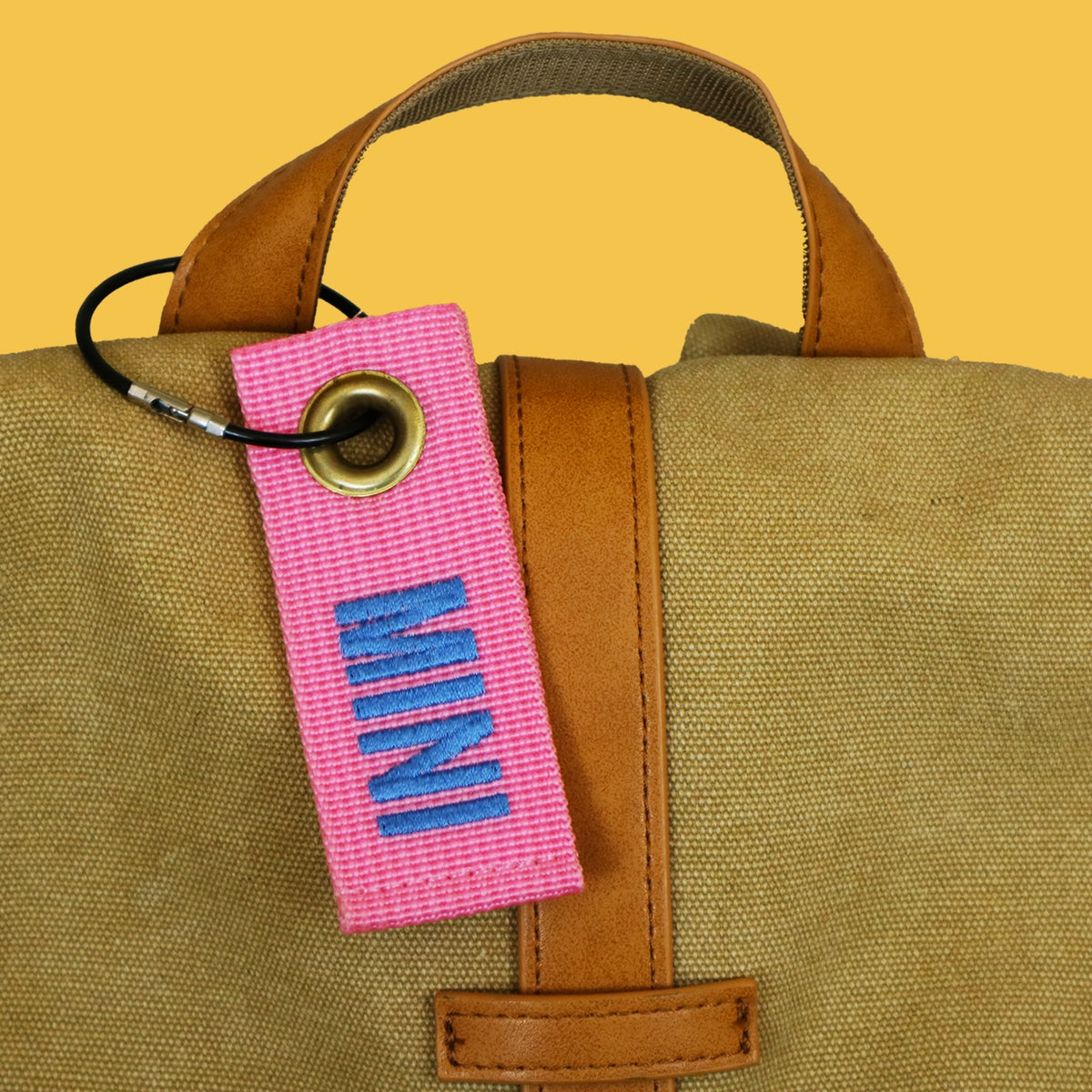 Pink mini bag tag embroidered MINI, hanging from a canvas bag handle on a black wire keyring