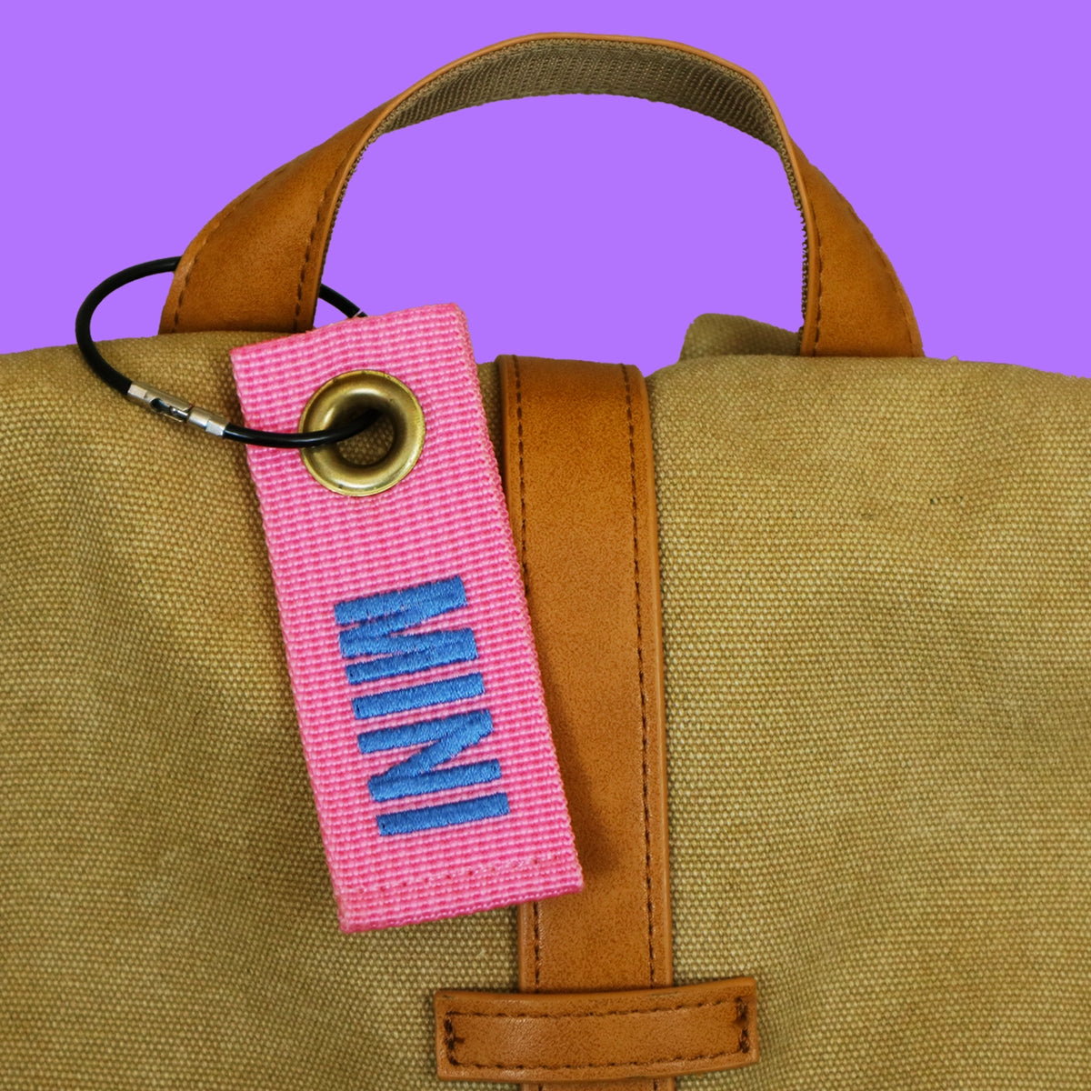 Pink mini bag tag embroidered MINI, hanging from a canvas bag handle on a black wire keyring